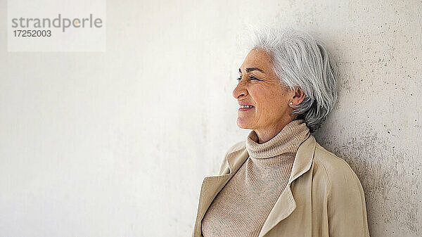 Lächelnde Frau mit kurzen Haaren träumt vor einer weißen Wand