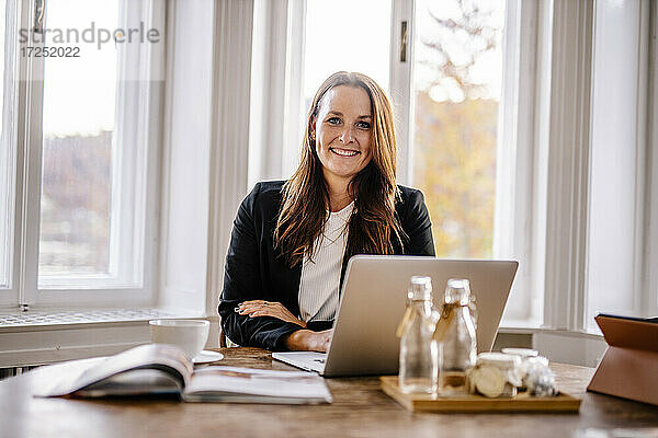 Lächelnde Veranstaltungsplanerin sitzt mit Laptop am Tisch im Büro
