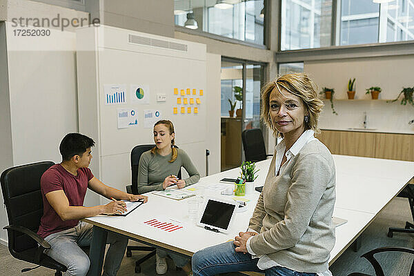 Geschäftsfrau sitzt auf dem Schreibtisch  während männliche und weibliche Mitarbeiter im Büro diskutieren