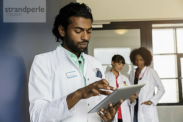 Junge männliche Mitarbeiter im Gesundheitswesen  die ein digitales Tablet benutzen  mit weiblichen Kollegen im Hintergrund im Krankenhaus