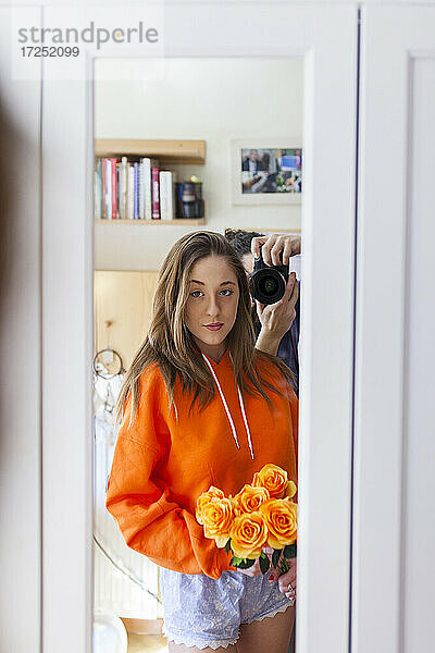 Junge Frau mit orangefarbenen Blumen schaut in den Spiegel  während ein Mann im Hintergrund durch die Kamera fotografiert