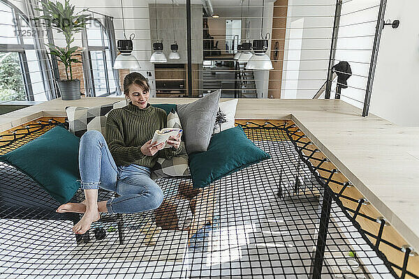 Eine Frau liest ein Buch  während sie auf einer Hängematte in einem modernen Haus sitzt