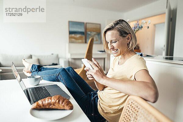 Lächelnde Geschäftsfrau mit hochgelegten Füßen  die bei der Arbeit zu Hause ein Mobiltelefon benutzt
