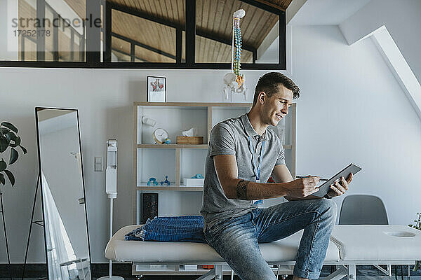 Männlicher Physiotherapeut  der ein digitales Tablet benutzt  während er auf einem Massagetisch in einer Arztpraxis sitzt