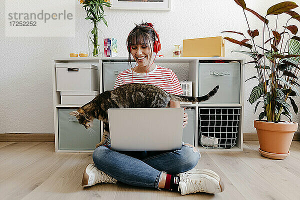 Junge Frau mit Kopfhörern sitzt mit Laptop und Katze zu Hause