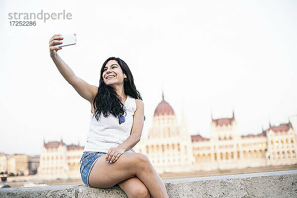 Schöne Frau  die ein Selfie mit ihrem Mobiltelefon macht  während sie auf einer Mauer sitzt  Budapest  Ungarn