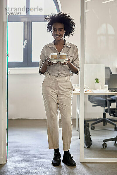 Afro-Geschäftsfrau  die Einweg-Kaffeebecher in der Hand hält  während sie in einem Büro an der Tür steht