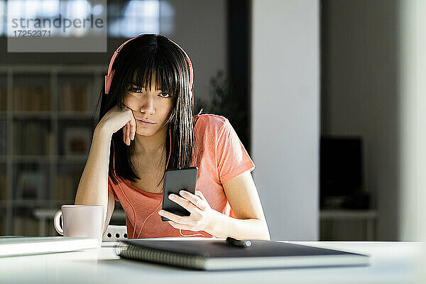 Frau mit Kopfhörern hält Smartphone  während sie zu Hause auf dem Tisch sitzt