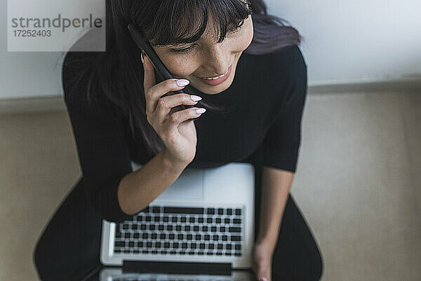 Mittlere erwachsene Frau  die mit dem Handy telefoniert  während sie zu Hause am Laptop sitzt