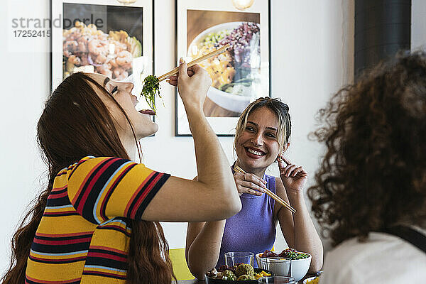 Lächelnde junge Frau sieht ihre Freundin an  die im Restaurant mit Stäbchen isst