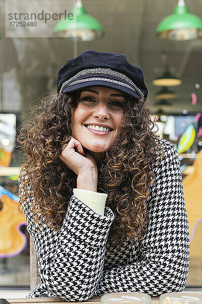 Lächelnde Frau mit lockigem Haar und Mütze sitzt mit Hand am Kinn in einem Straßencafé