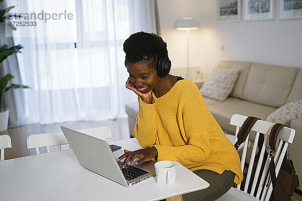 Lächelnde junge Frau  die einen Laptop benutzt  während sie zu Hause über Kopfhörer Musik hört