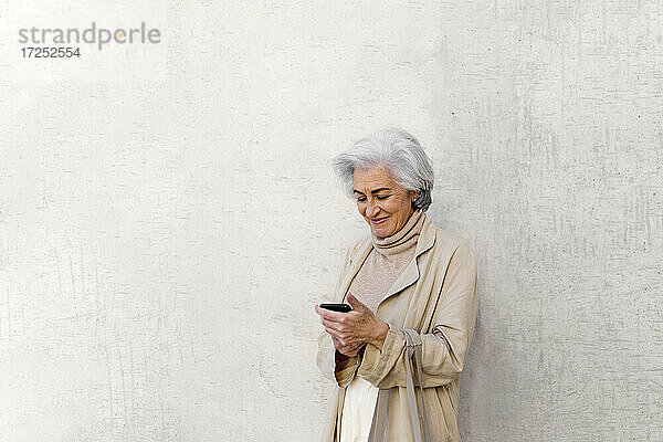 Lächelnde reife Frau  die ein Mobiltelefon vor einer Wand benutzt