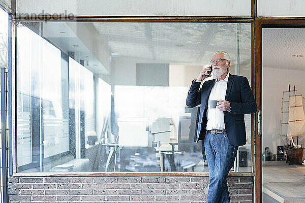 Geschäftsmann mit Kaffeetasse  der mit seinem Smartphone telefoniert und sich an eine Glaswand im Büro lehnt