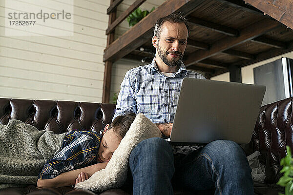 Mann benutzt Laptop  während sein Sohn zu Hause auf dem Sofa schläft