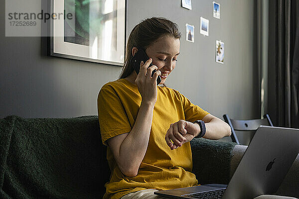 Lächelnde Frau  die die Zeit überprüft  während sie zu Hause mit ihrem Handy telefoniert