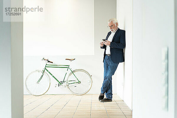 Ein älterer männlicher Unternehmer benutzt ein Smartphone  während er sich im Büro an die Wand lehnt