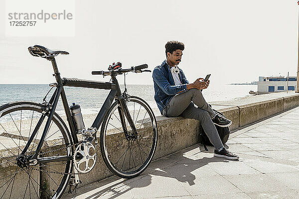 Mann  der ein Mobiltelefon benutzt  während er an einem sonnigen Tag mit dem Fahrrad auf einer Stützmauer sitzt