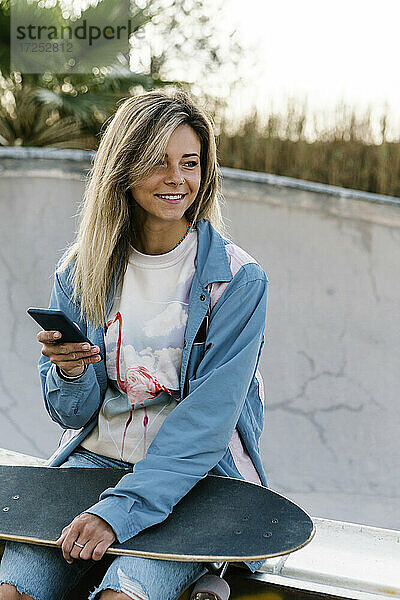 Lächelnde Frau mit Skateboard  die wegschaut  während sie ein Handy im Park hält