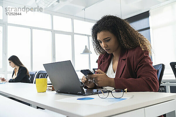 Geschäftsfrau  die ein Mobiltelefon benutzt  während im Hintergrund eine Kollegin in einem Coworking-Büro arbeitet