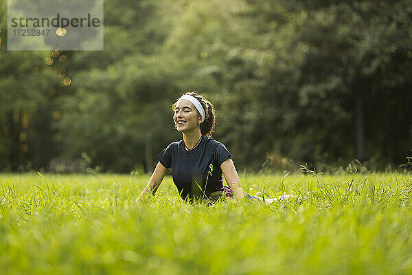 Lächelnde Frau übt Yoga auf Gras in einem öffentlichen Park
