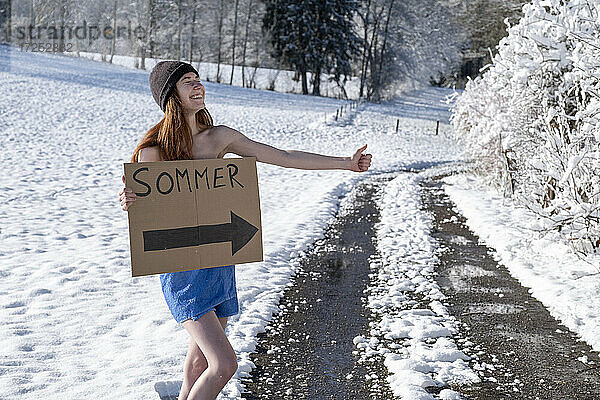 Lächelnde Frau mit Schild  die auf einer schneebedeckten Straße trampt