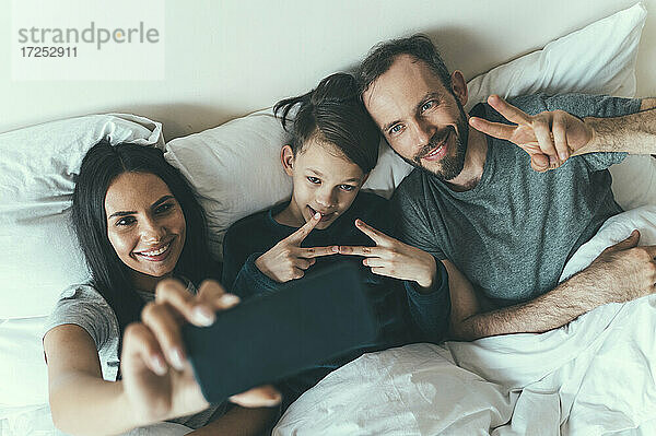 Vater und Sohn gestikulieren  während die Frau ein Selfie auf dem Bett liegend zu Hause macht
