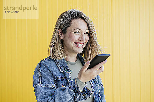 Glückliche Frau  die eine Sprachnachricht über ihr Smartphone an eine gelbe Wand sendet