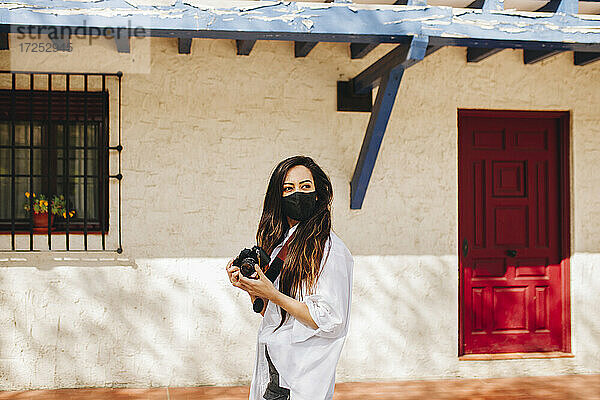 Junge Touristin mit Gesichtsschutzmaske  die eine Kamera hält  während sie an einem Haus in einem Dorf steht