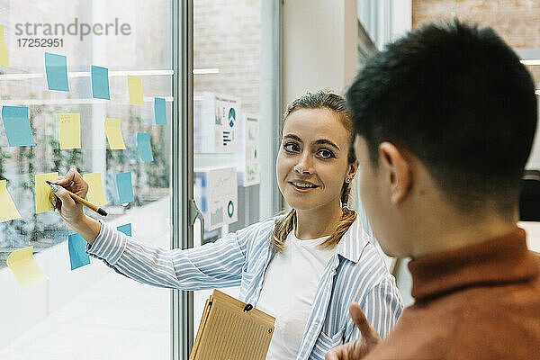 Lächelnde Unternehmerinnen und Unternehmer  die über Haftnotizen an der Glaswand eines Coworking-Büros diskutieren