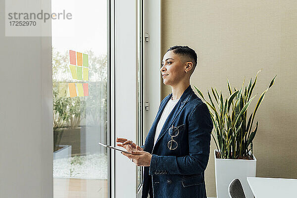 Nachdenkliche Geschäftsfrau mit digitalem Tablet am Fenster im Büro stehend