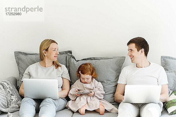 Lächelnde männliche und weibliche Unternehmer mit Laptop schauen sich an  während sie neben ihrer Tochter auf dem Sofa im Wohnzimmer sitzen