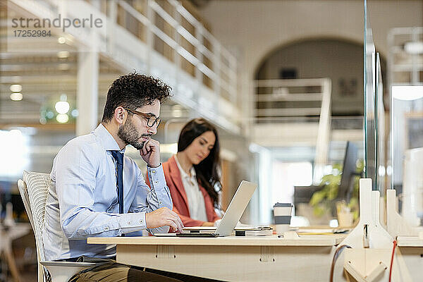 Männlicher Fachmann mit Hand am Kinn  der an einem Laptop arbeitet  mit einer weiblichen Kollegin im Hintergrund in einem Coworking-Büro