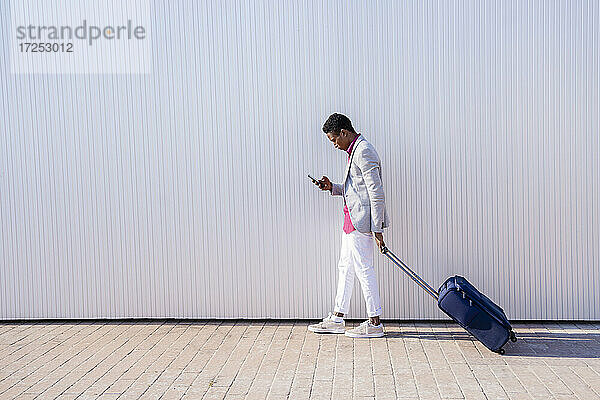 Geschäftsmann  der einen Koffer zieht  während er auf dem Gehweg sein Smartphone benutzt