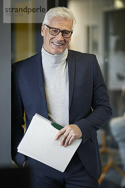 Lächelnder Unternehmer mit Dokument und Hand in der Tasche im Büro stehend