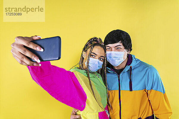 Junge Frau mit Gesichtsmaske nimmt Selfie durch Handy mit Freund vor gelbem Hintergrund