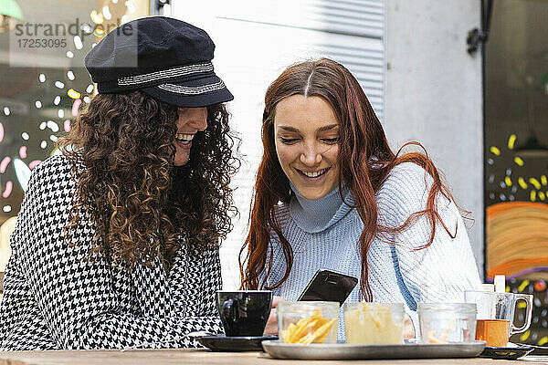 Lächelnde junge Frauen  die in einem Straßencafé ein Mobiltelefon benutzen