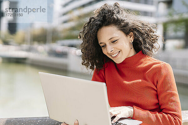 Lächelnde junge Frau  die an einem sonnigen Tag einen Laptop am Geländer benutzt