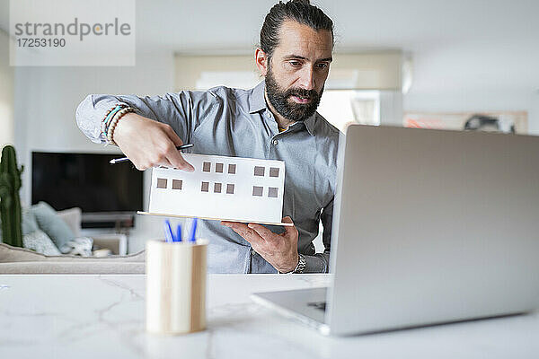 Männlicher Architekt  der ein architektonisches Modell hält  während er zu Hause einen Videoanruf tätigt
