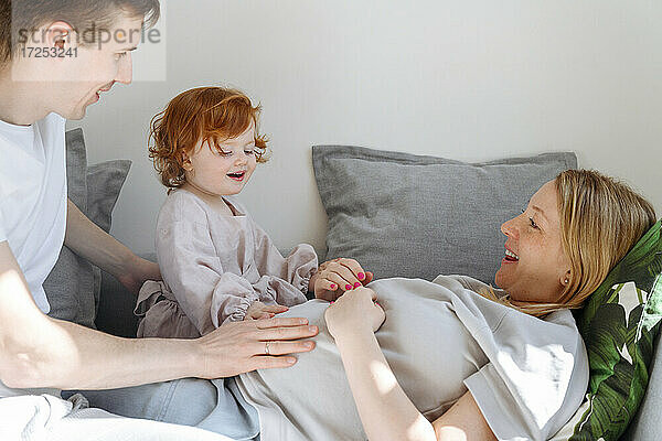 Lächelnder Vater und Tochter berühren den Bauch der schwangeren Frau auf dem Sofa zu Hause