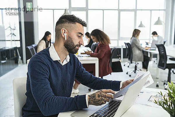 Ein männlicher Unternehmer liest ein Dokument  während er vor einem Laptop sitzt und mit männlichen und weiblichen Kollegen in einem Coworking-Büro arbeitet.