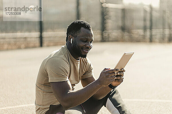Lächelnder gut aussehender Mann  der ein digitales Tablet benutzt  während er an einem sonnigen Tag auf einem Sportplatz sitzt