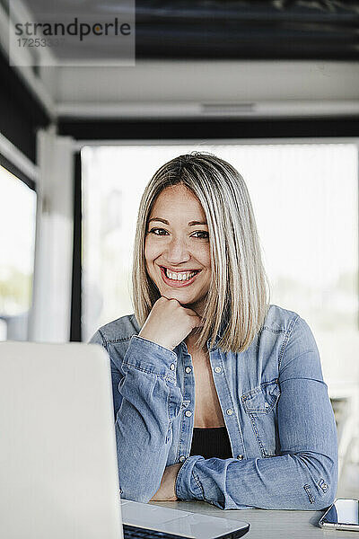 Glückliche Geschäftsfrau mit Hand am Kinn  die mit einem Laptop in einem Café sitzt