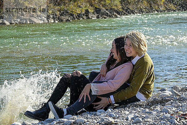 Fröhliches lesbisches Paar  das am Seeufer sitzt  während das Wasser an einem sonnigen Tag auf die Steine spritzt