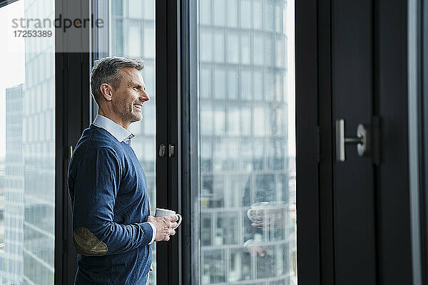 Lächelnder reifer Geschäftsmann mit Kaffeetasse  der durch ein Fenster im Büro schaut