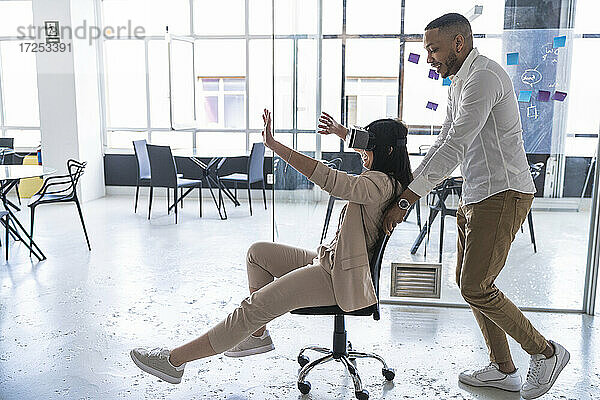 Männliche Fachkraft schiebt Geschäftsfrau  die einen Virtual-Reality-Simulator auf einem Stuhl in einem Coworking-Büro trägt