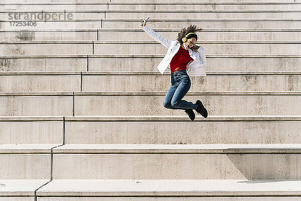 Ekstatische Frau mit Kopfhörern und Smartphone springt an einem sonnigen Tag auf einer Treppe