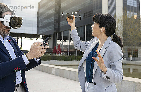 Geschäftsmann mit Virtual-Reality-Headset  der eine Drohne fernsteuert  während er mit einer Kollegin im Büropark steht