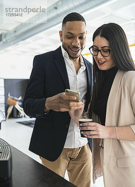 Lächelnde männliche und weibliche Fachkräfte  die während der Kaffeepause in einem Coworking-Büro ein Mobiltelefon benutzen