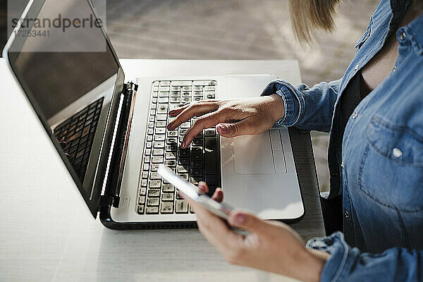 Unternehmerin hält ihr Handy in der Hand  während sie in einem Café am Laptop arbeitet